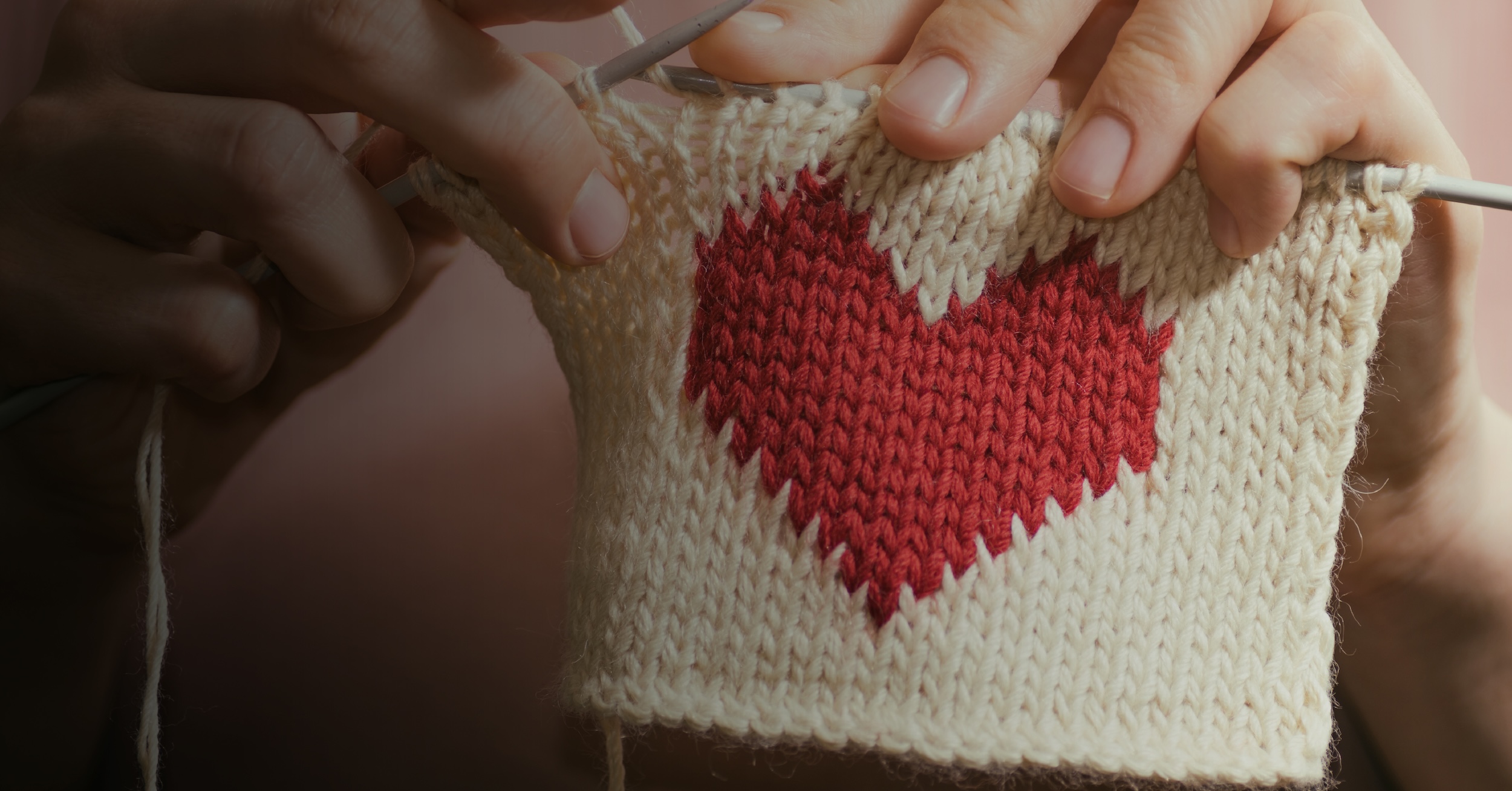 Hands knitting a cream fabric swatch with a red heart pattern.