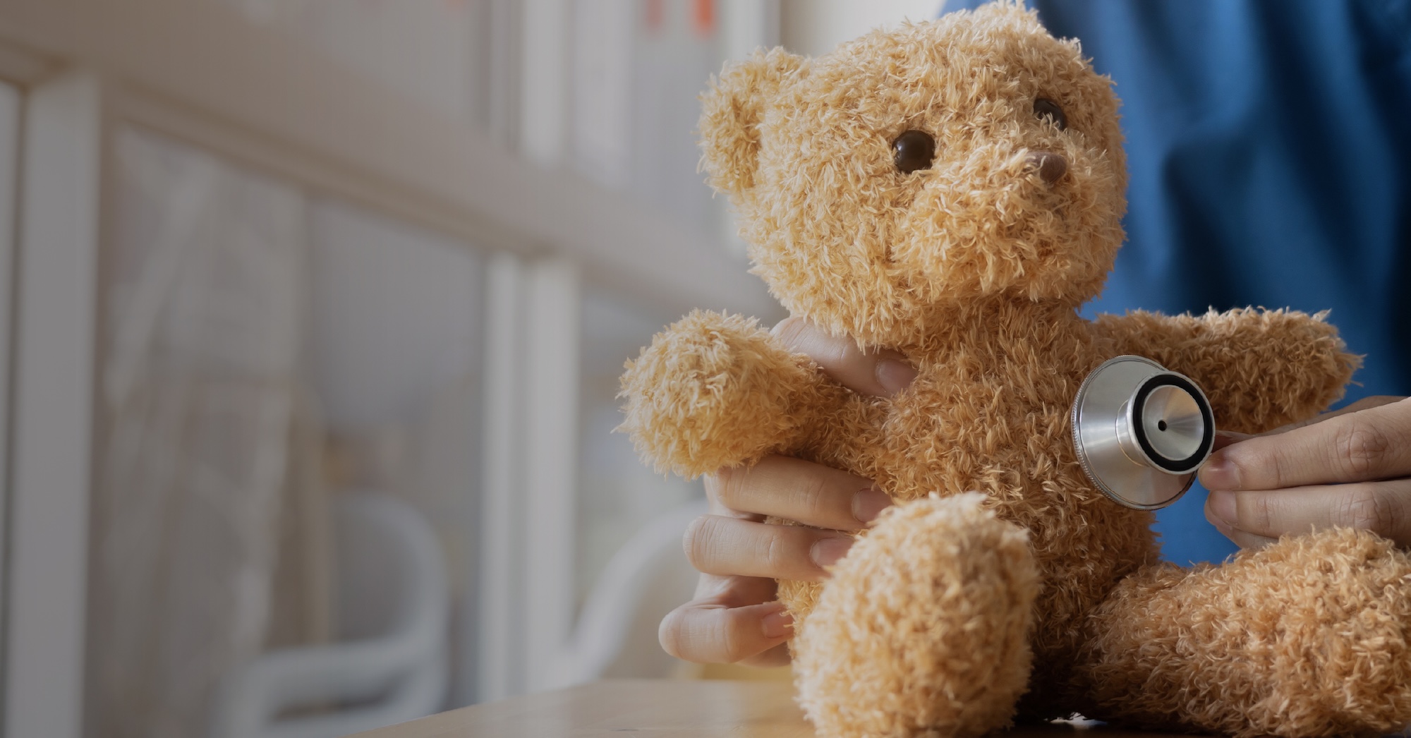 Doctor holding a teddy bear and listening to its chest with a stethoscope in a pediatric exam room.