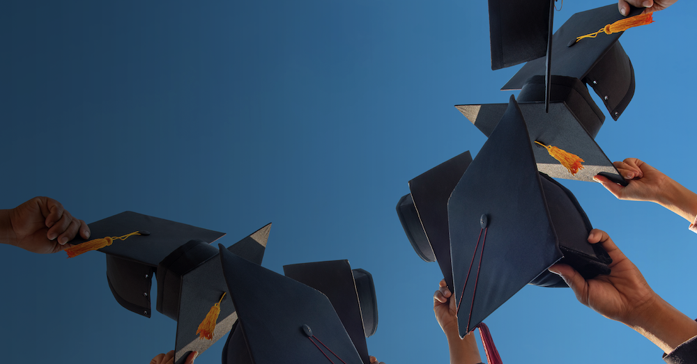 Graduates hold black graduation caps with gold tassels against a blue sky.
